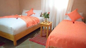 A room with two beds, each with an orange blanket and pillow, a small wooden table with a vase of flowers, and a window with sheer curtains. - Riad Essaouira (Essaouira)
