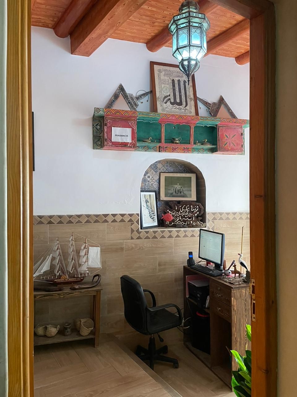 A room with a wooden ceiling, a desk with a computer, a chair, a shelf with decorative items, and a small ship model.