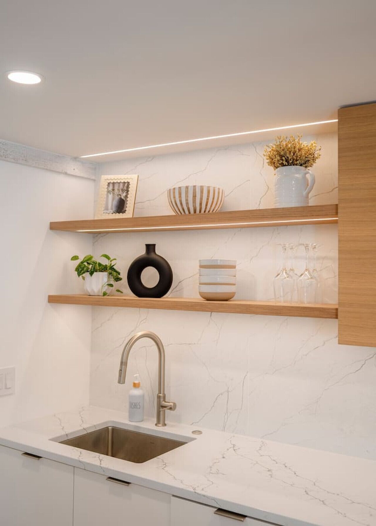 A modern kitchen with a marble countertop, a stainless steel sink, wooden shelves, and decorative items.