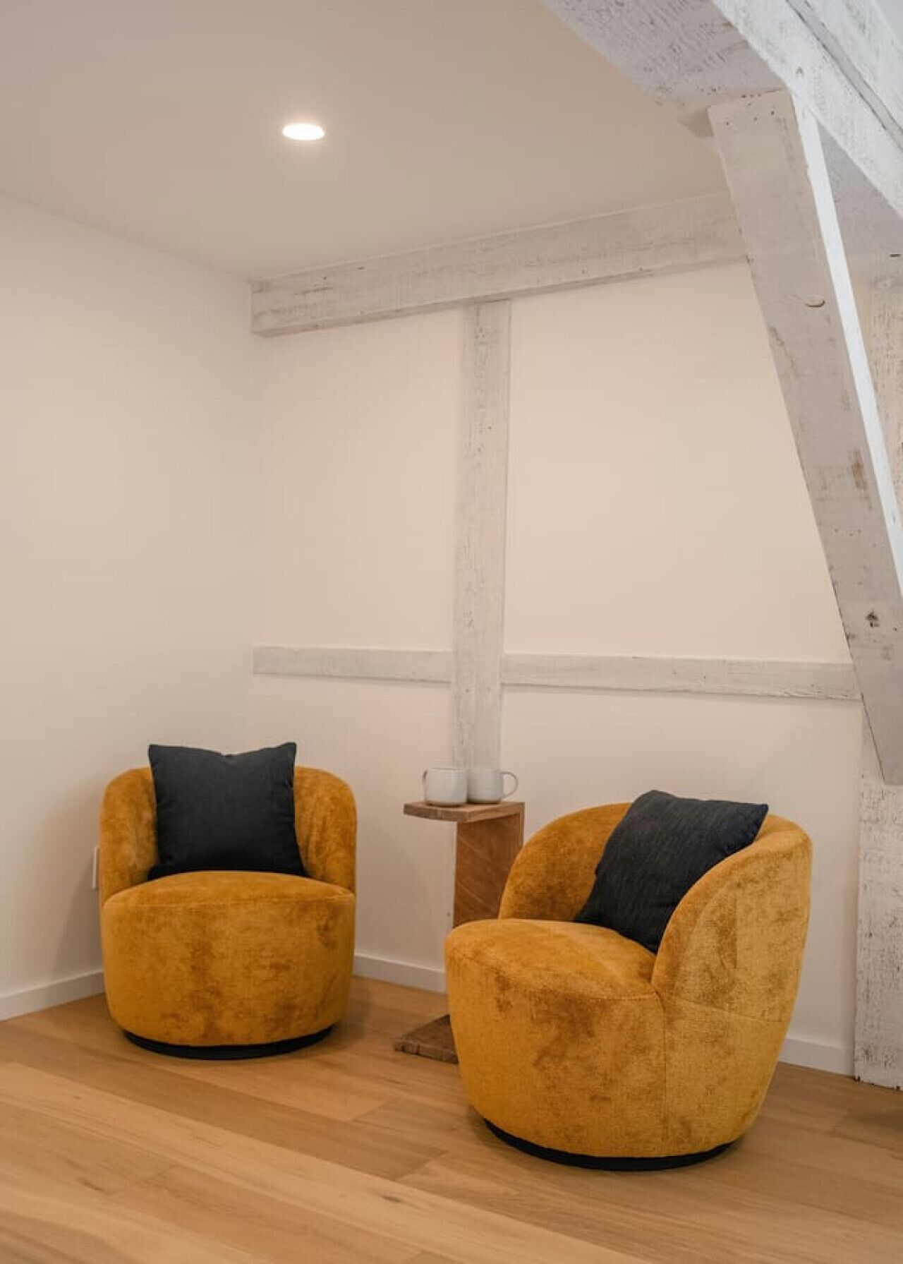 Two mustard-colored armchairs with dark cushions, a small wooden table with two white cups, and a white wall with exposed wooden beams.
