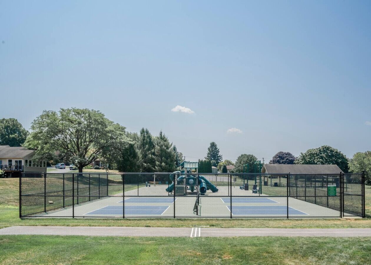 Ein eingezäunter Außenbereich mit Spielplatz, einschließlich Rutschen und einer Kletterkonstruktion, umgeben von Bäumen und blauem Himmel.