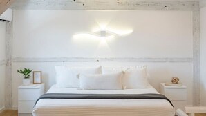 A minimalist bedroom with a large bed, white walls, exposed wooden beams, and a modern light fixture.