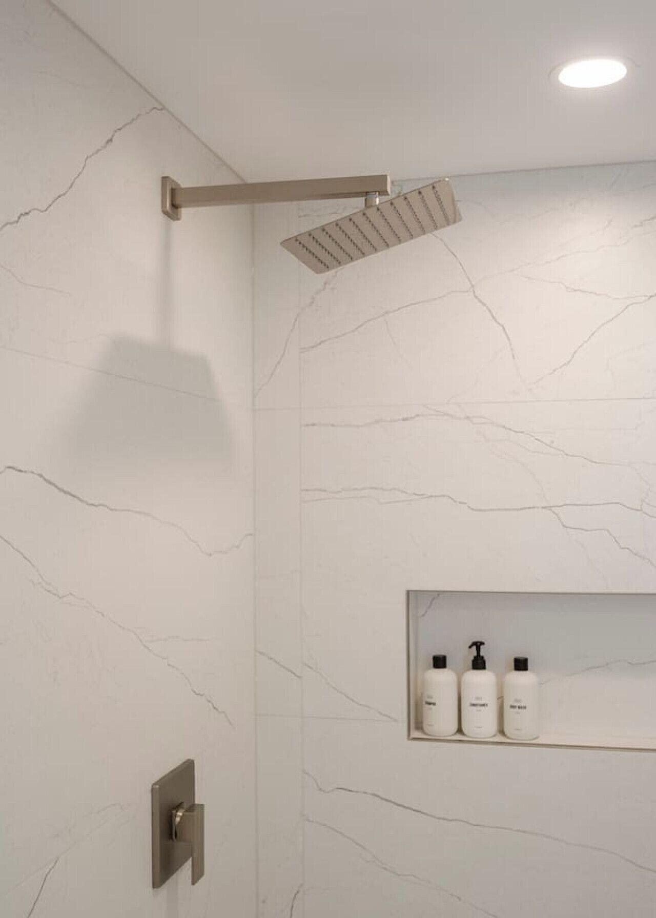 A modern bathroom with a marble wall, a shower with a horizontal rain showerhead, and a shelf with three bottles of toiletries.