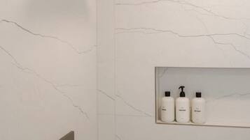 A modern bathroom with a marble wall, a shower with a horizontal rain showerhead, and a shelf with three bottles of toiletries.