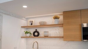A modern kitchen with white marble countertops, a stainless steel sink, wooden shelves with decorative items, and built-in wooden cabinets.