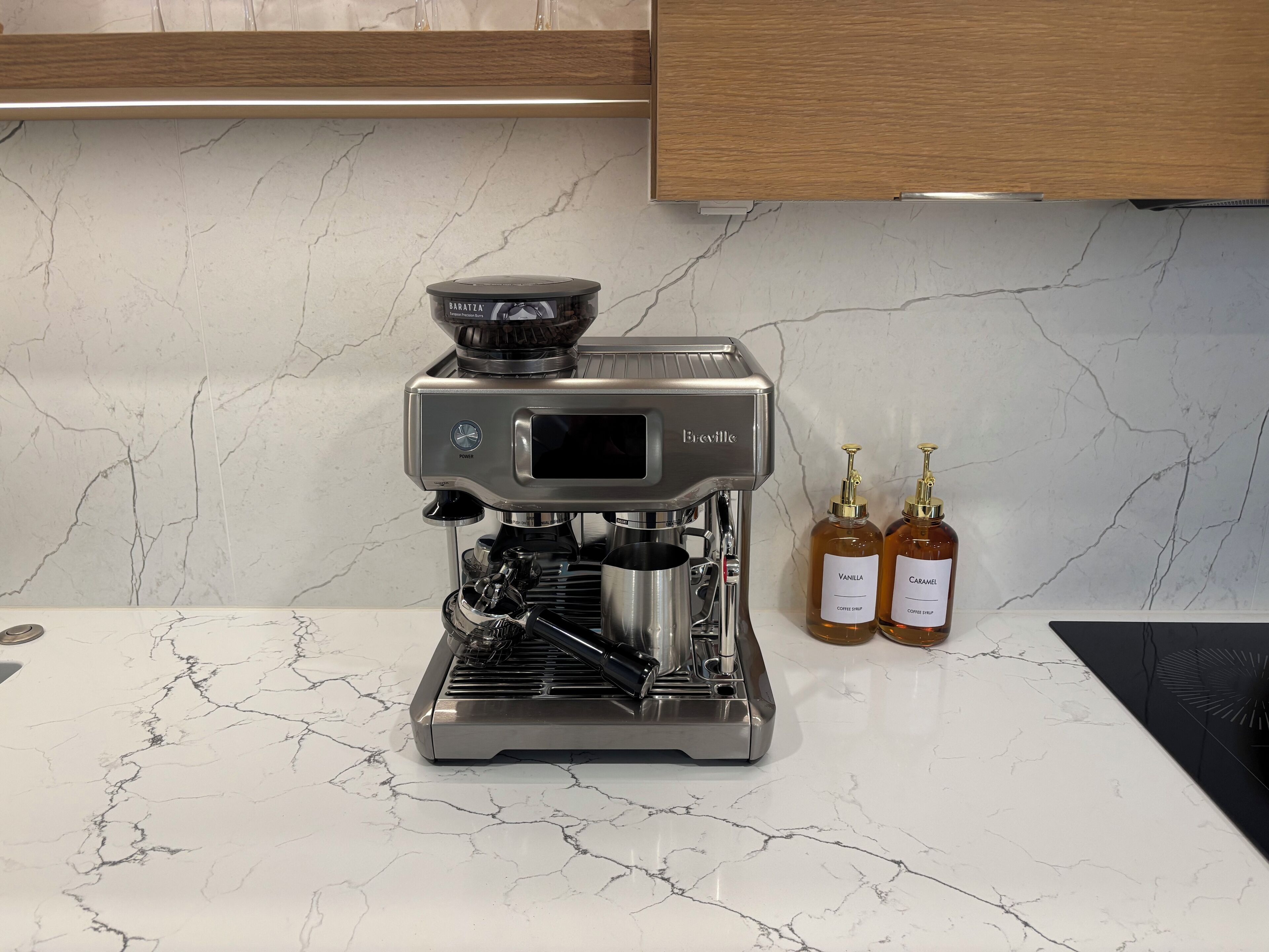 A Breville espresso machine on a marble countertop with two coffee syrup bottles labeled "Vanilla" and "Caramel."