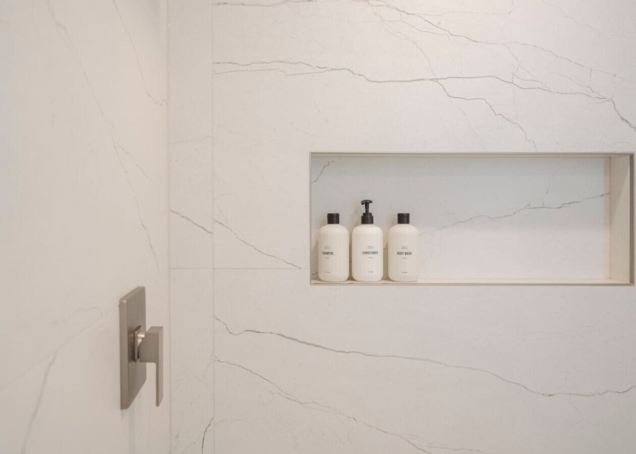 Un baño con pared de mármol, un estante empotrado con tres frascos de artículos de aseo, y un grifo moderno.