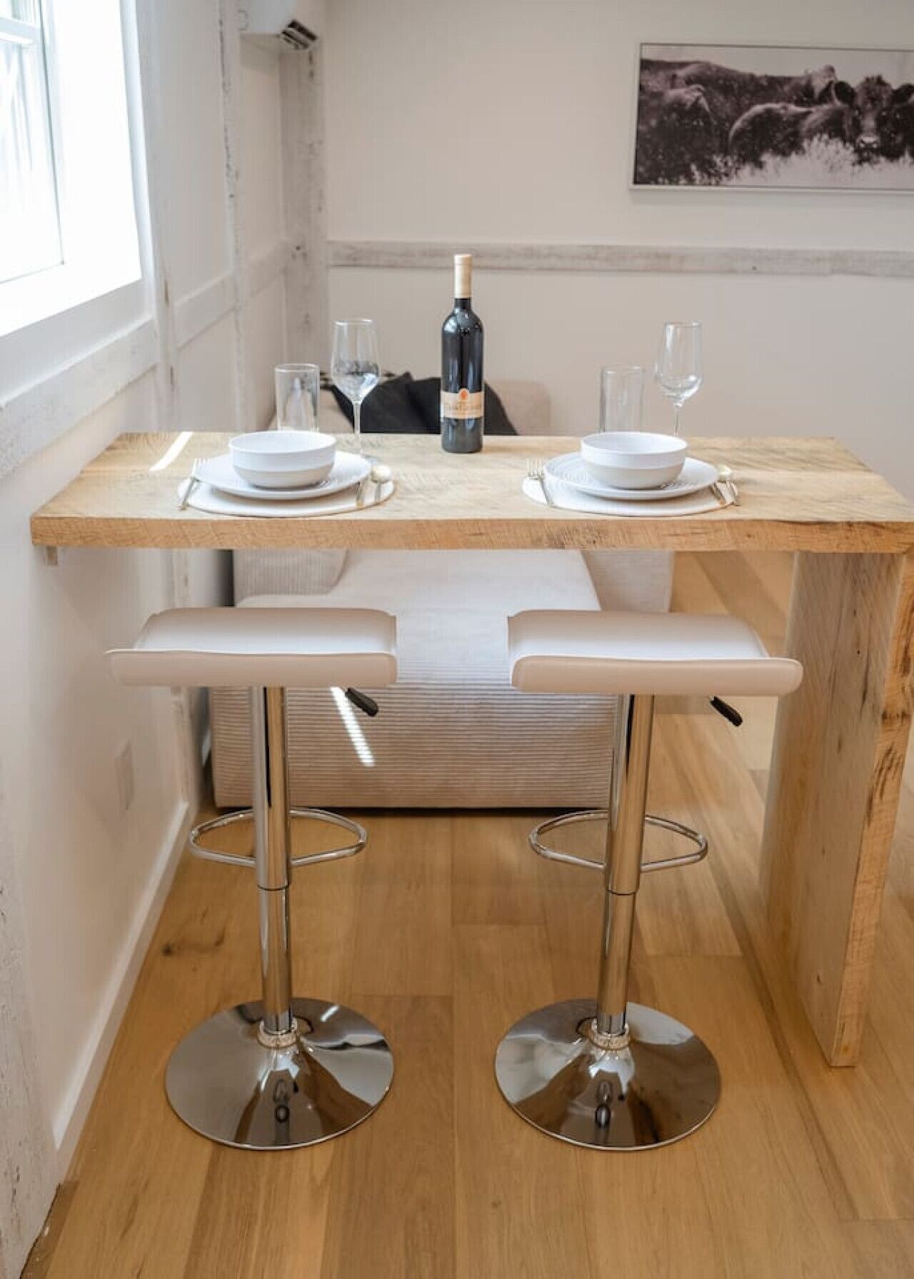 A small dining area with a wooden table, two white barstools, and a bottle of wine.