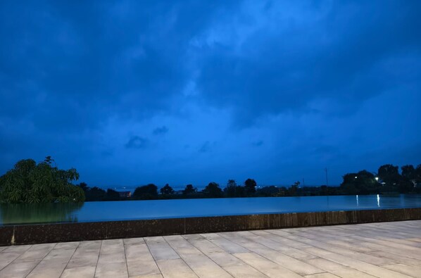A waterfront view at dusk with a calm body of water, a wooden deck, and distant trees. - River's Edge By Escape Stays (Nashik)