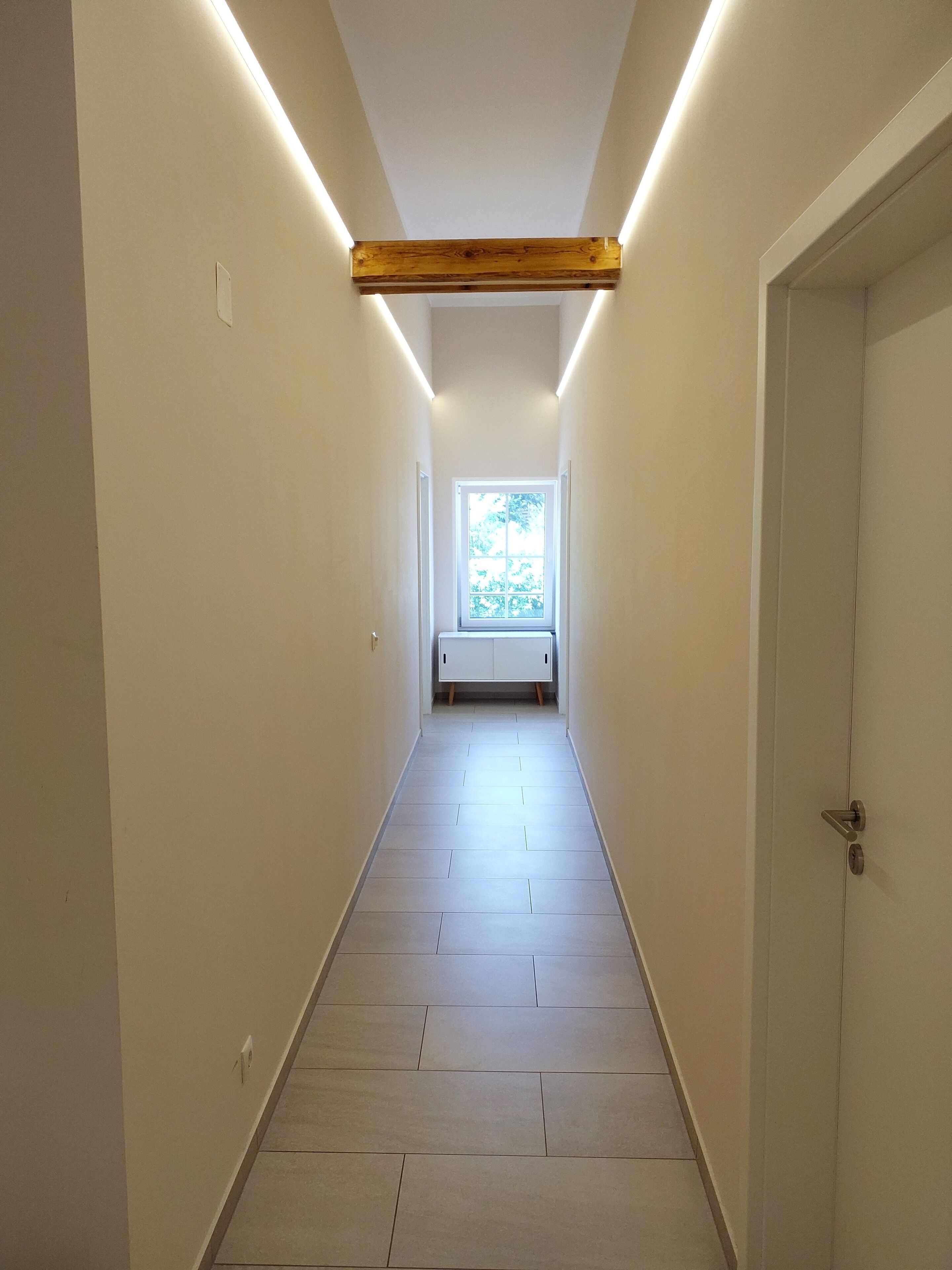 A narrow hallway with a wooden beam ceiling, a window at the end, and a white door on the right.