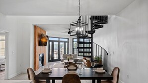 Uma sala de jantar moderna com uma mesa de madeira escura, cadeiras brancas e uma escada em espiral preta ao fundo.