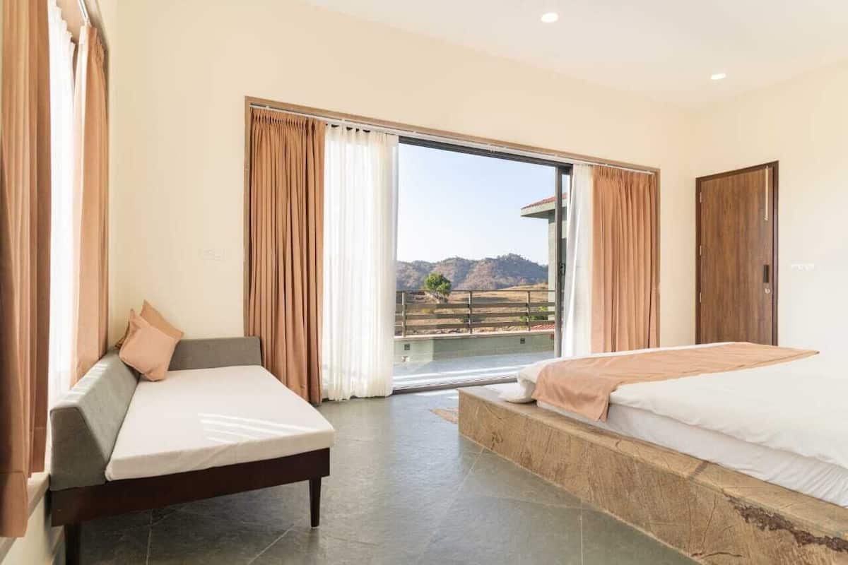 A hotel room with a bed, a sofa, and a large sliding glass door leading to a balcony with a mountain view.