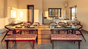 Een eetruimte met een lange tafel, meerdere stoelen en bankjes. Achtergrond toont een keuken met een raam.