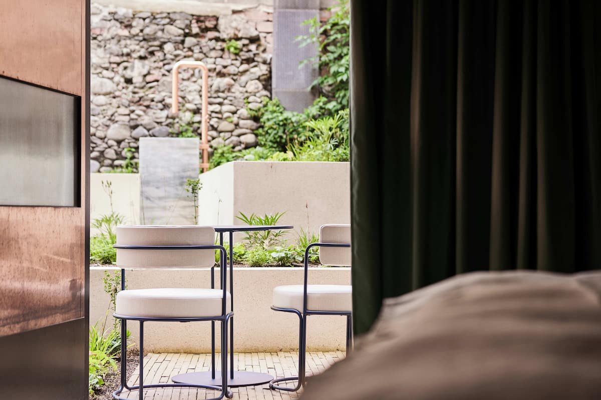Habitación de hotel con vista a un área exterior con mobiliario, una pared de piedra, plantas y una silla de metal.