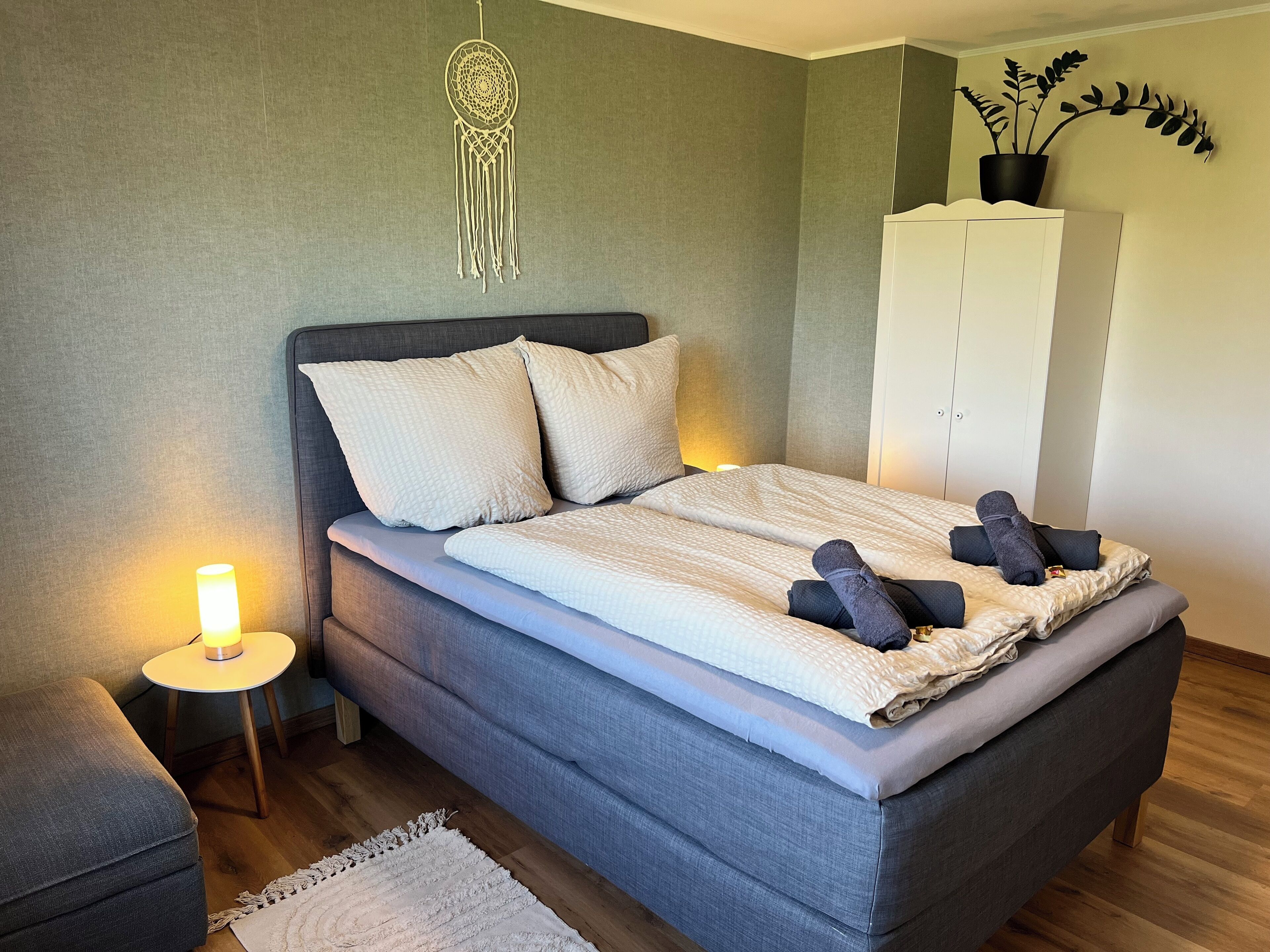 Una cama bien hecha con almohadas y una colcha, una lámpara de noche, una mesita con lámpara, un sofá gris, una alfombra blanca y un armario.