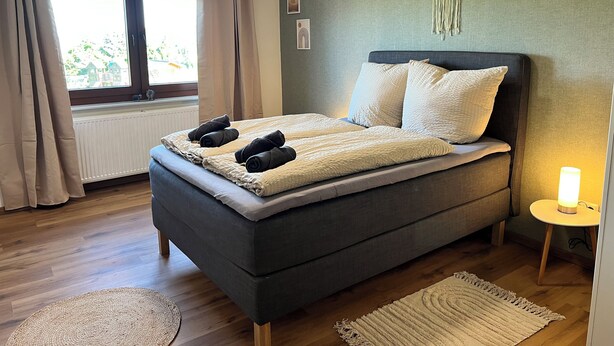 Ein modernes Schlafzimmer mit einem großen Bett, einem Nachttisch mit Lampe und Blick ins Freie durch ein Fenster.