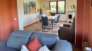 Ein modernes Wohnzimmer mit einem grauen Sofa, einem Essbereich mit Tisch und Stühlen sowie einem großen Fenster mit Jalousien.