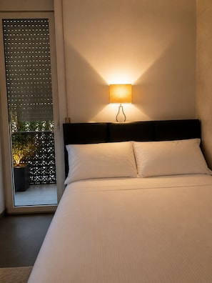 A bedroom with a bed, a wall-mounted lamp, and a view through a window with blinds.