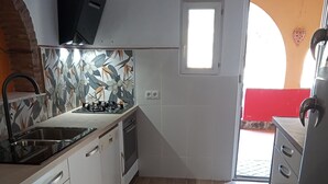 Een compacte keuken met witte kasten, een gasfornuis en een spoelbak. Er is een klein raam en een deur naar buiten.