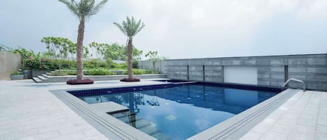 A rectangular swimming pool with a tiled edge, surrounded by a paved deck. There are two palm trees and a small bush in the foreground.