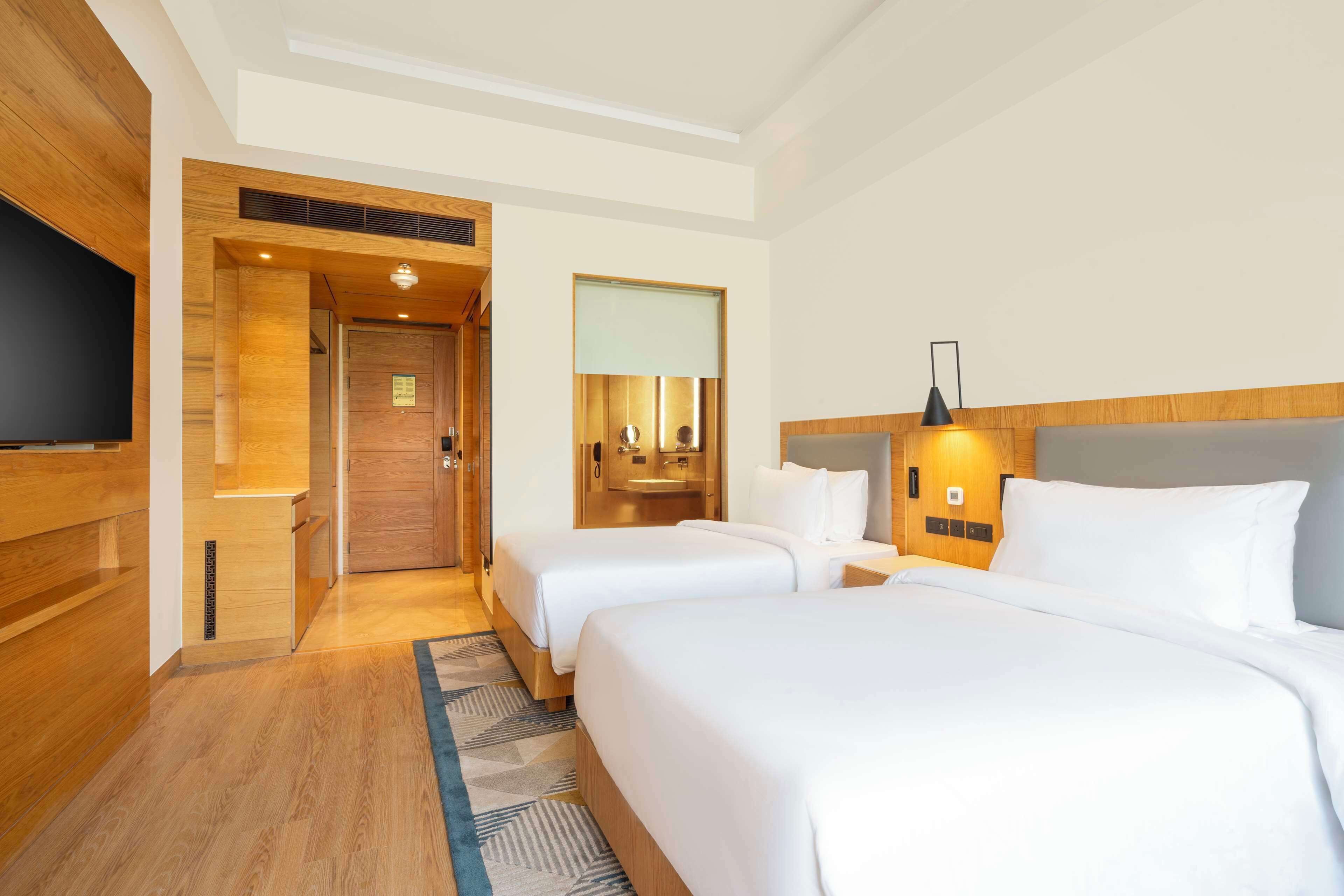 A hotel room with two beds, a television, a bathroom with a mirror, and a wooden floor.
