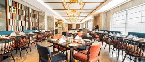 A spacious dining area with multiple tables and chairs, a central chandelier, and a view of the kitchen in the background.
