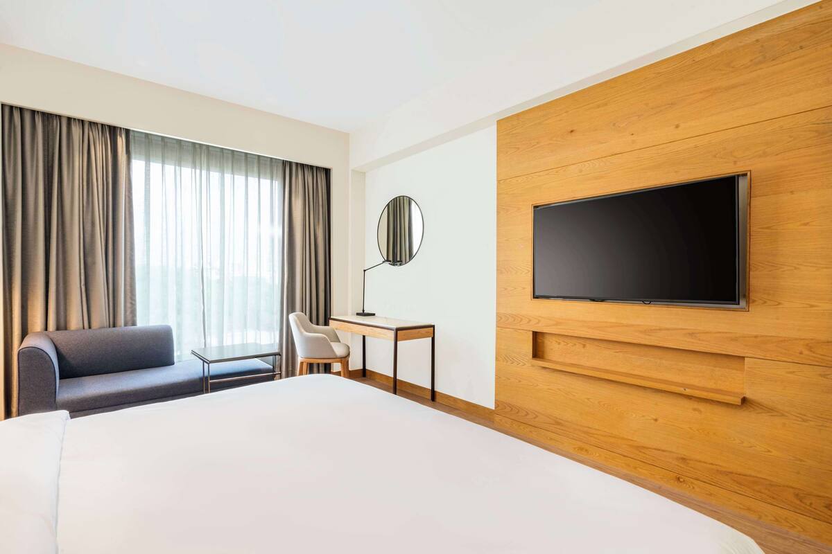 Une chambre d’hôtel avec un grand lit, un bureau, une chaise, un téléviseur fixé à un mur en bois et une fenêtre avec des rideaux.