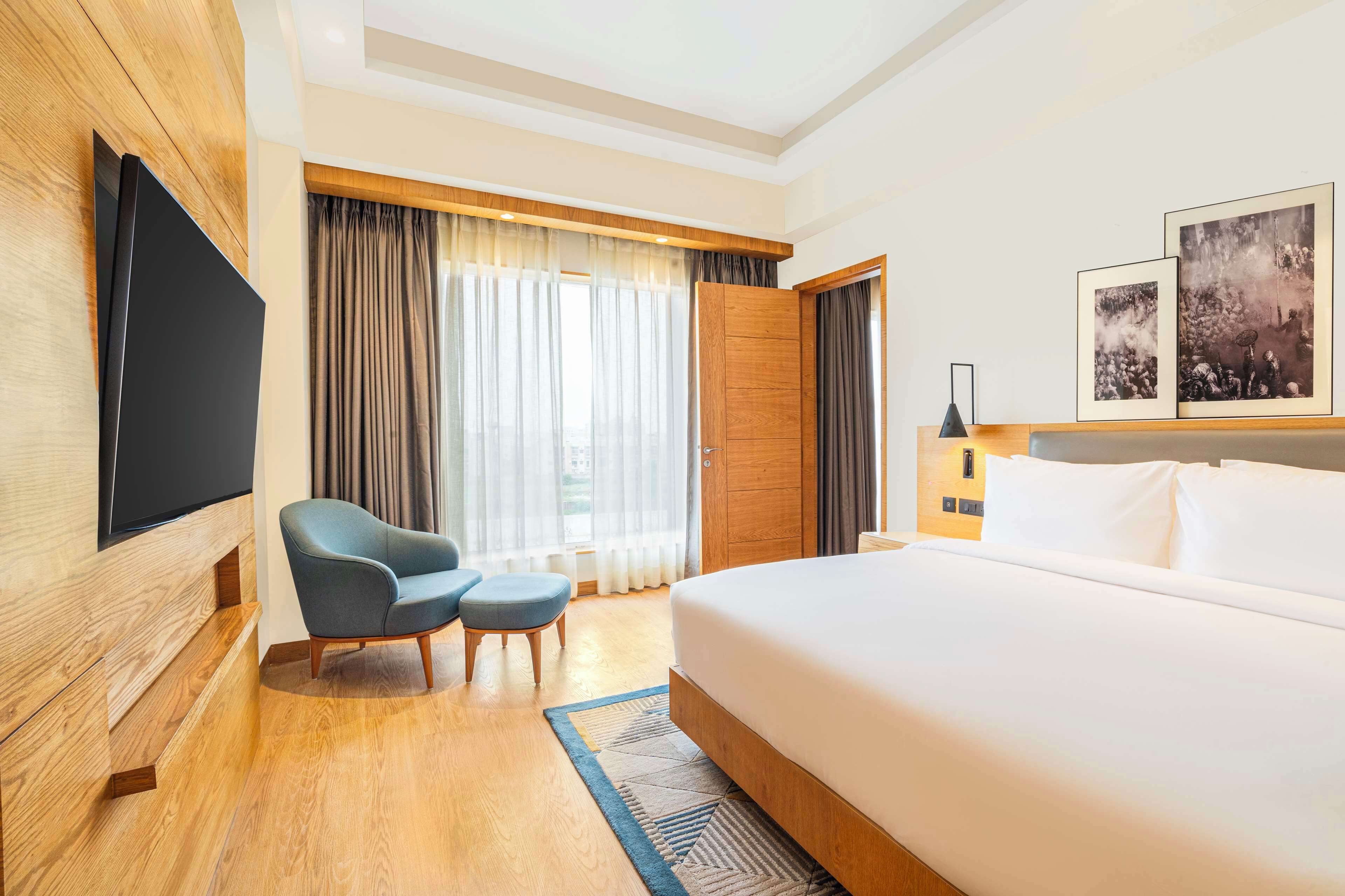 A hotel room with a large bed, a TV mounted on the wall, a seating area with a chair and ottoman, and a window with curtains.