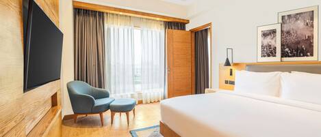 Een hotelkamer met een groot bed, een aan de muur bevestigde televisie, een zithoek met een fauteuil en poef, en een raam met gordijnen.
