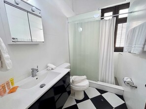 Une salle de bain avec un lavabo, des toilettes et une douche, le tout de couleur blanche. Un miroir est placé au-dessus du lavabo et il y a une fenêtre avec des rideaux.