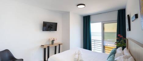 Une chambre d’hôtel avec un lit, une chaise, une télévision, une fenêtre avec des rideaux et une vue sur la mer.