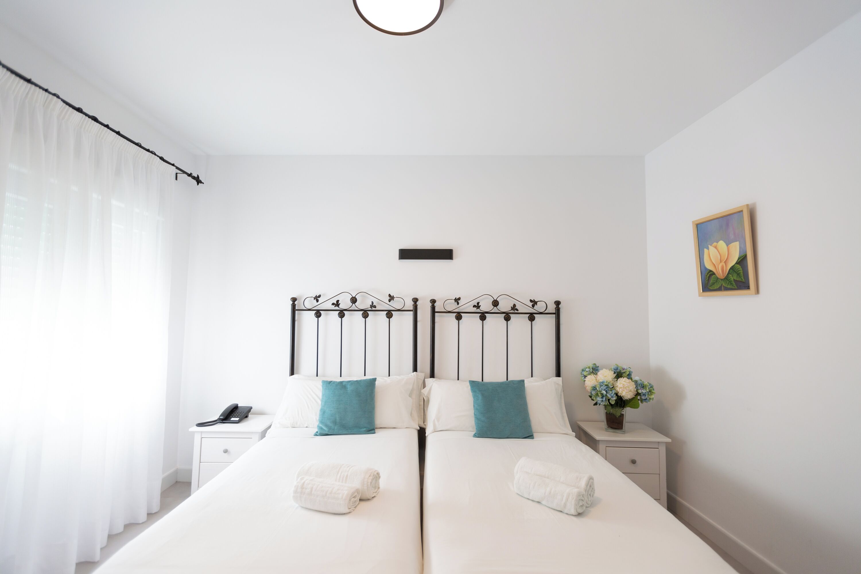 Een tweepersoonsbed met witte lakens en teal kussens, een nachtkastje met een telefoon, en een bloemenvaas op het nachtkastje.