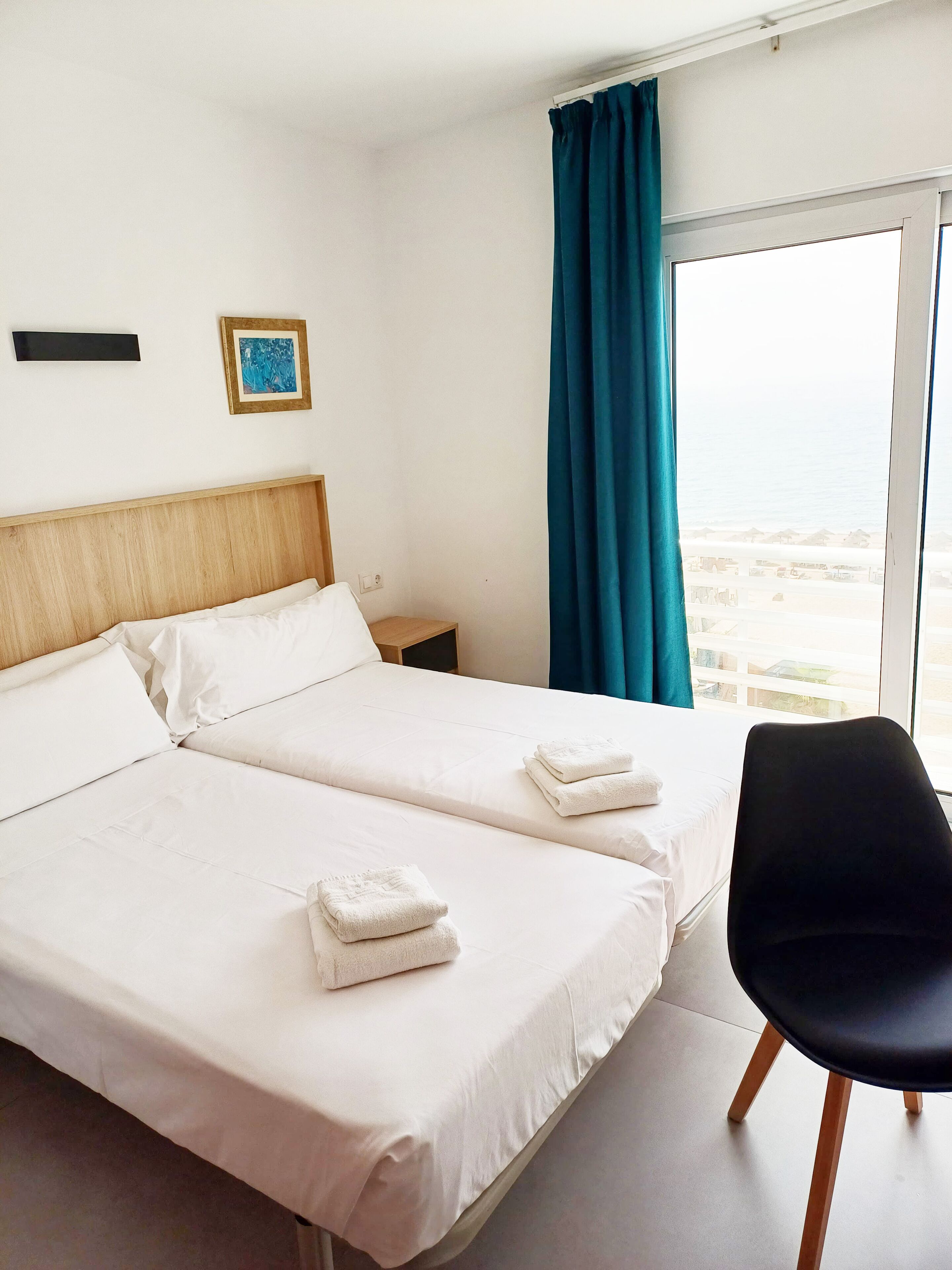 A hotel room with two beds, a chair, a nightstand, a picture on the wall, and a view of the beach through the window.