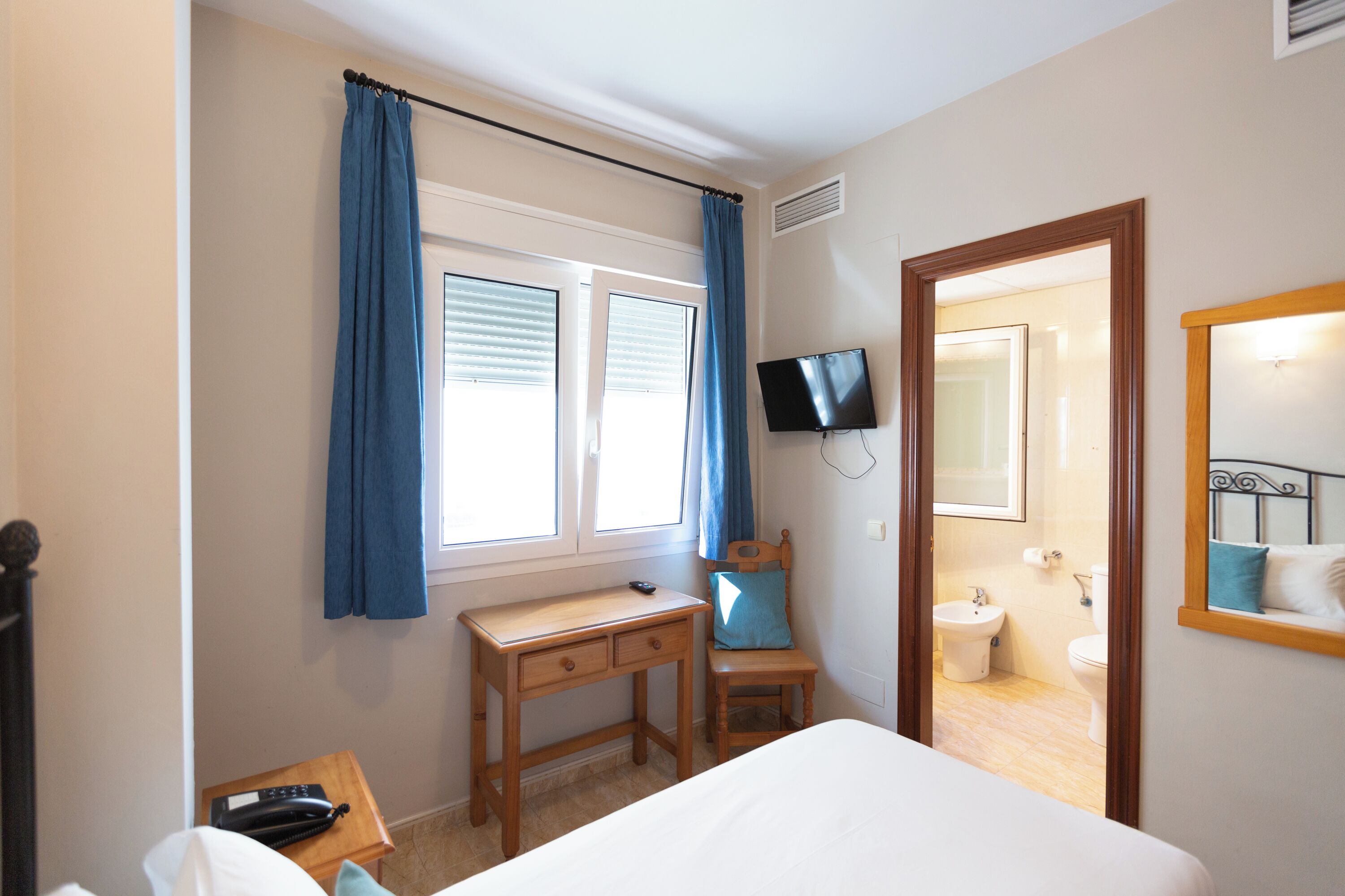 Ein Schlafzimmer mit Bett, Schreibtisch, Stuhl, Fernseher und einem durch eine offene Tür einsehbaren Badezimmer.