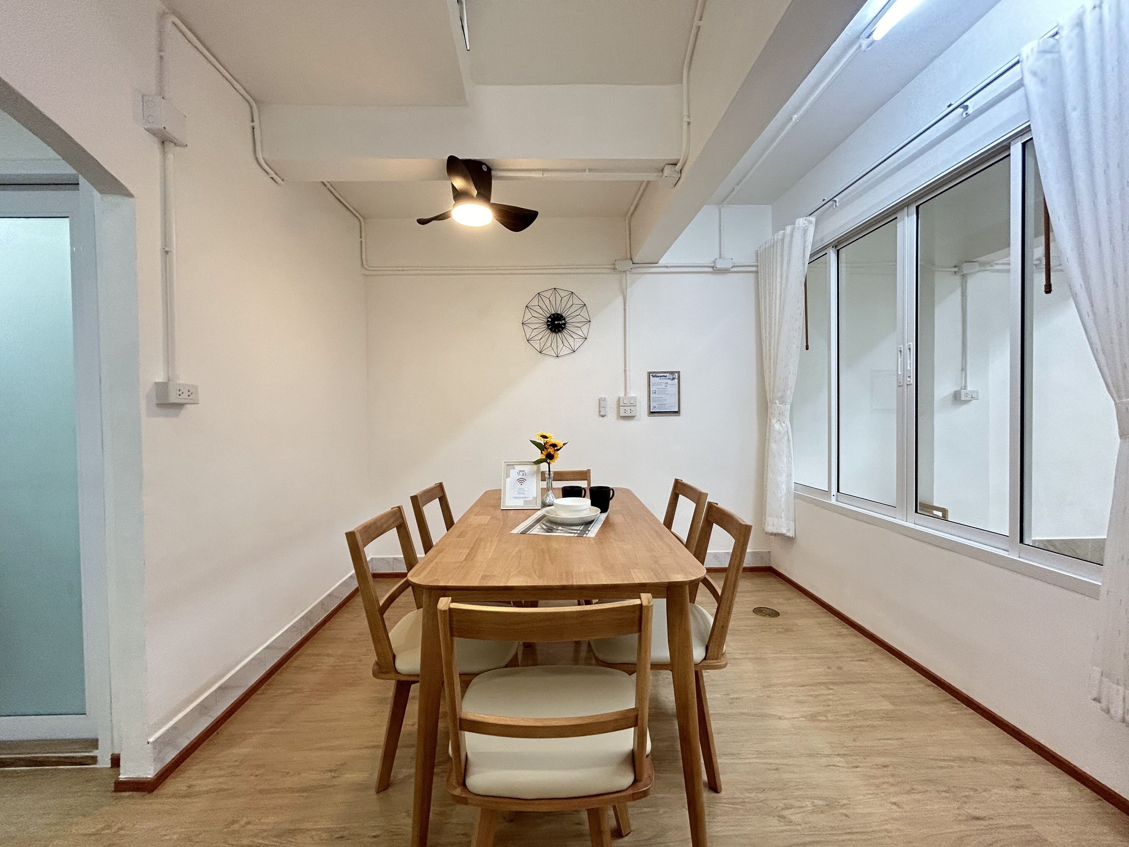 A dining area with a wooden table, six chairs, a ceiling fan, a decorative wall piece, and a large window with curtains.