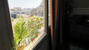 Et værelse med et vindue der giver udsigt over bybilledet, en potteplante med røde blomster og udsigt til bygninger.