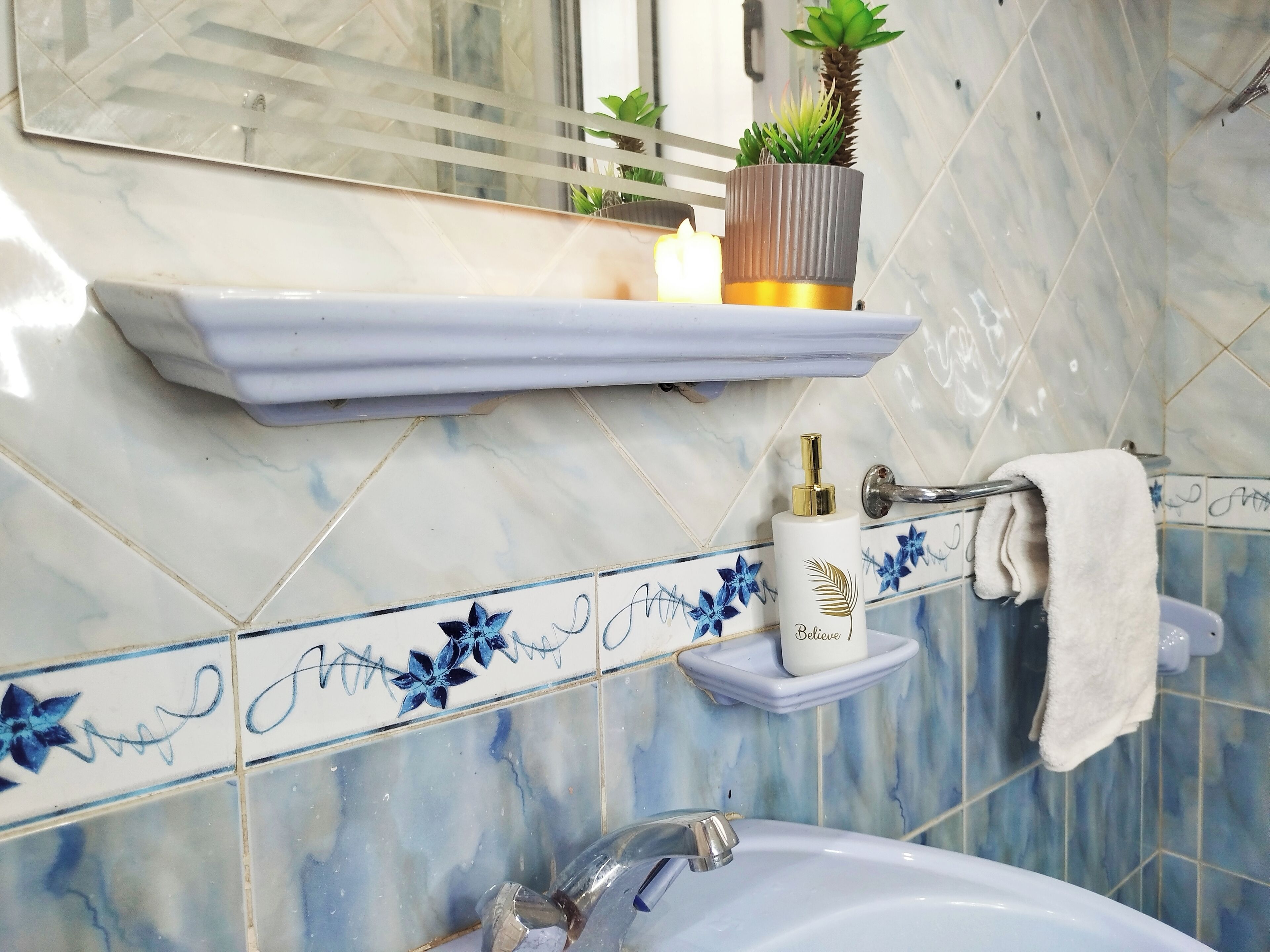 A bathroom with a marble backsplash, a white sink, a mirror, a shelf with a plant and a candle, and a towel hanging on a rack.
