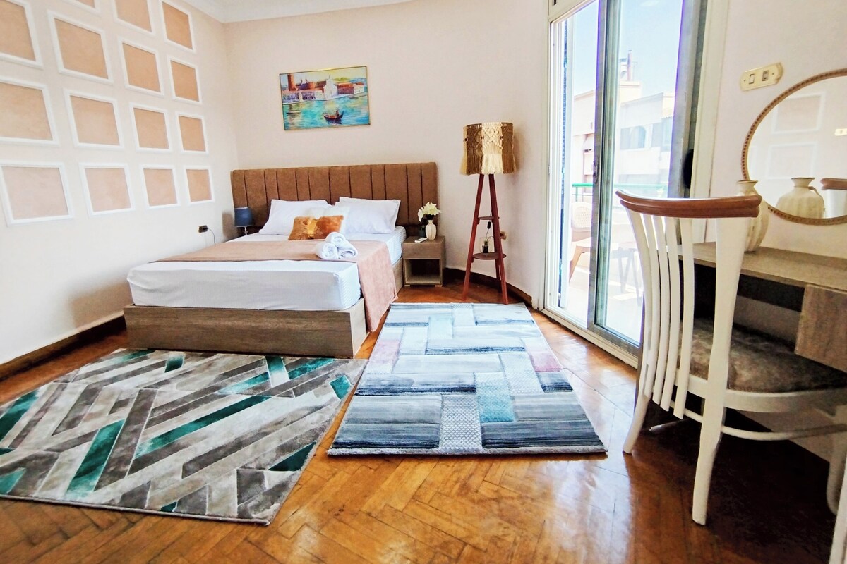 A hotel room with a bed, two bedside tables, a nightstand, a lamp, a chair, a window, and a painting on the wall.