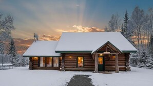 Une cabane en rondins dont le toit est recouvert de neige, avec un porche à l’avant et une cheminée bien visible.
