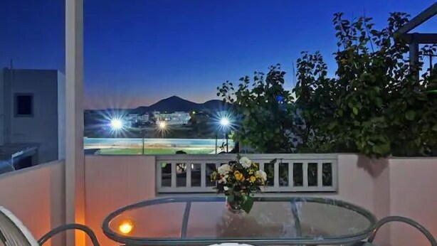 A balcony with a glass table and chairs, a vase with flowers, and a view of a lit sports field and mountains at dusk. - Memories of Milos (Milos)