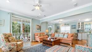 A spacious living room with a large sofa, a coffee table, and a patterned ottoman. There is a ceiling fan, a window with blinds, and a door leading outside. - 4BR Home w/ Free Linens & Beach Access (Surfside Beach)