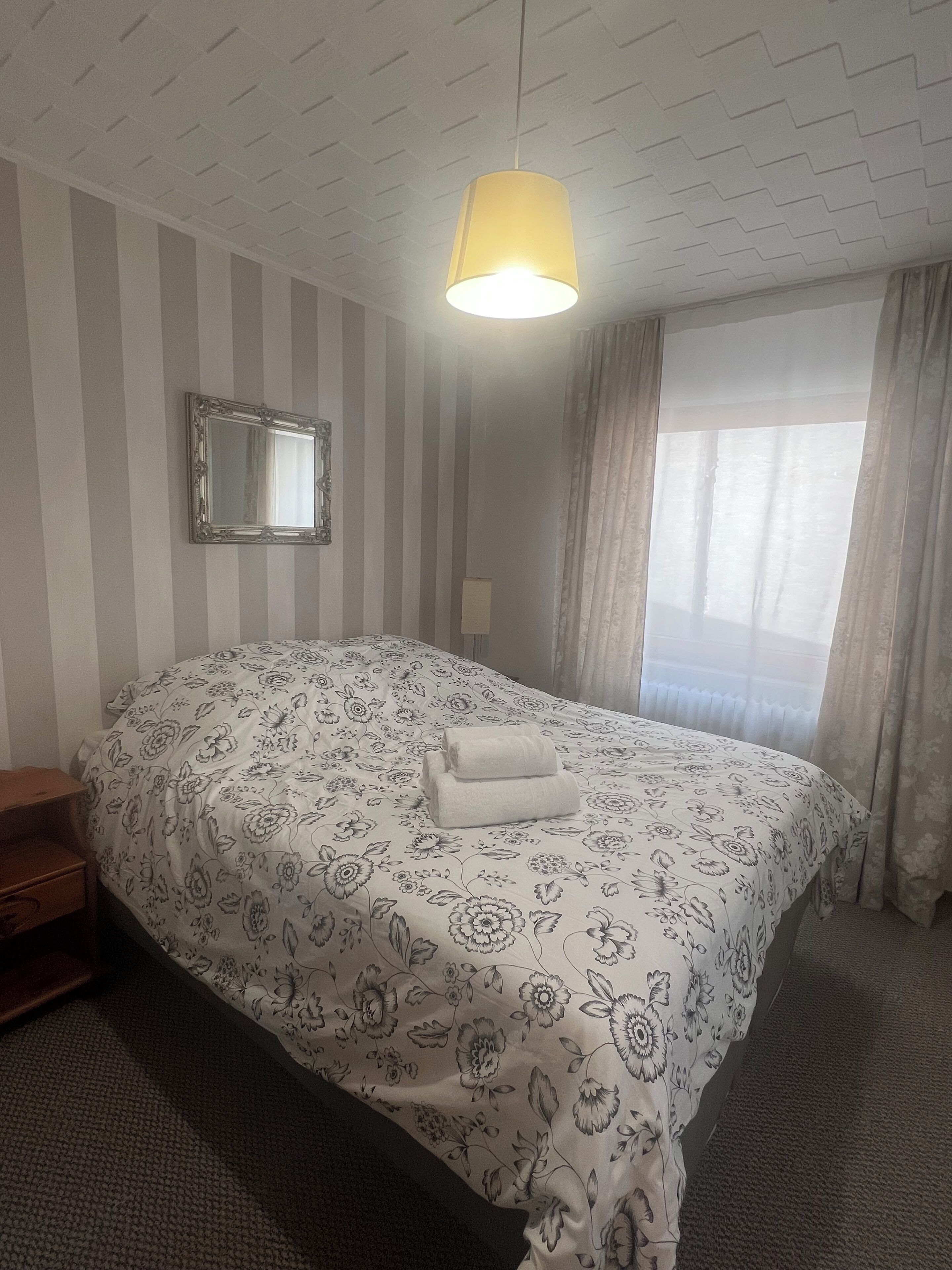 A hotel room with a bed, a bedside table, a mirror, and a window with curtains.