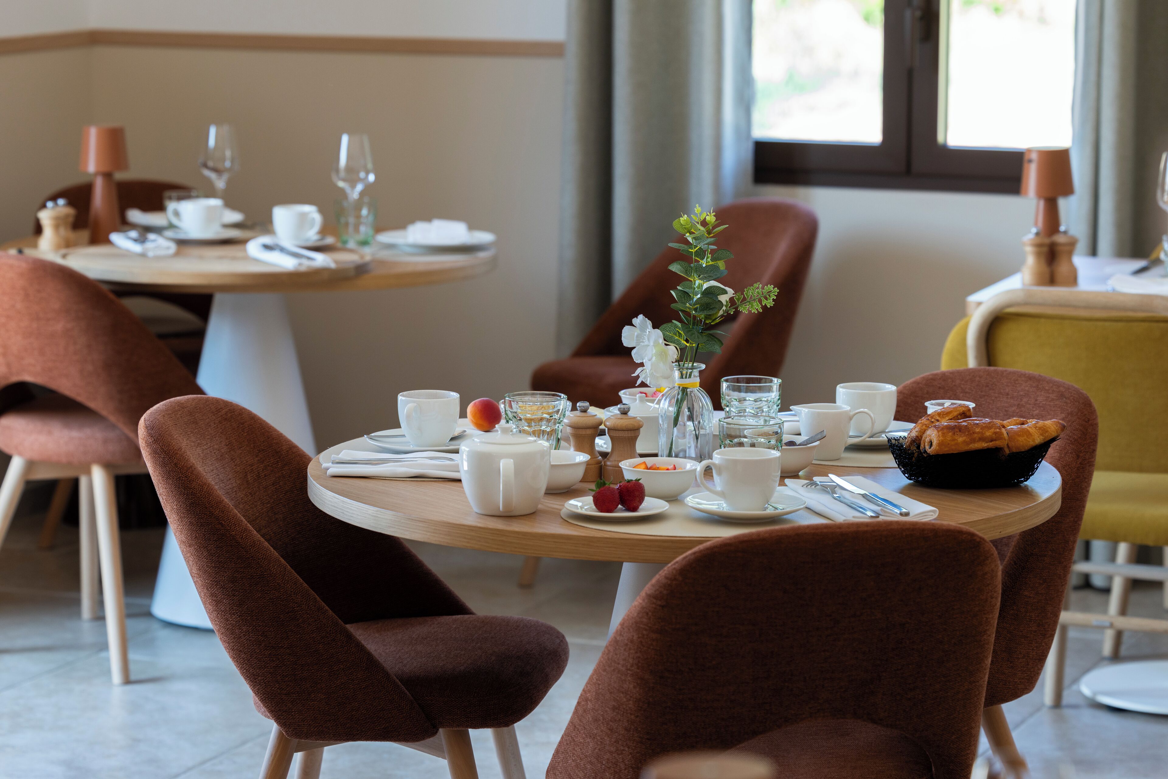 A dining area with round tables, brown upholstered chairs, and a variety of table settings including plates, cups, and a vase with flowers.