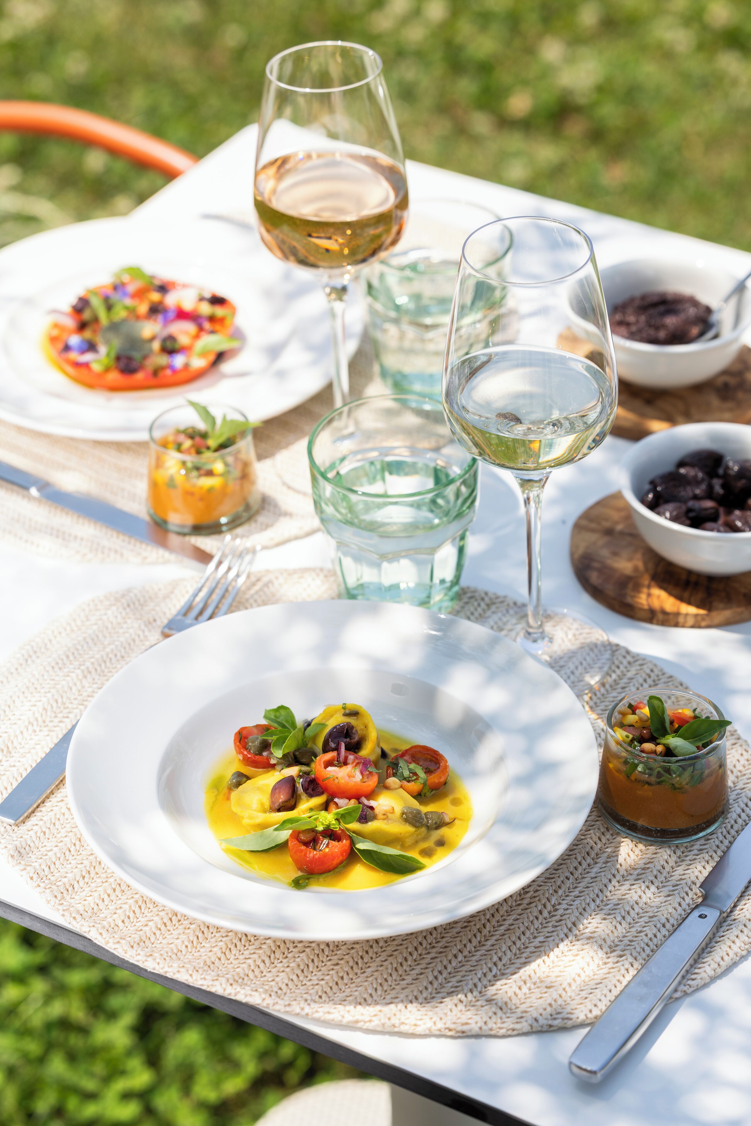 A table set outdoors with plates of food, glasses of wine, and small bowls of olives and olives.