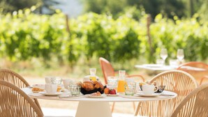 A table set for breakfast with croissants, fruit, and juice, surrounded by wicker chairs in an outdoor setting with a vineyard in the background.