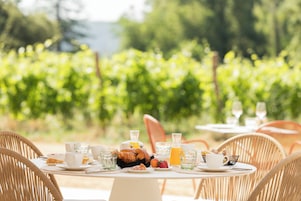 Una mesa dispuesta para el desayuno con cruasanes, fruta y jugo, rodeada de sillas de mimbre en un entorno al aire libre con un viñedo de fondo.