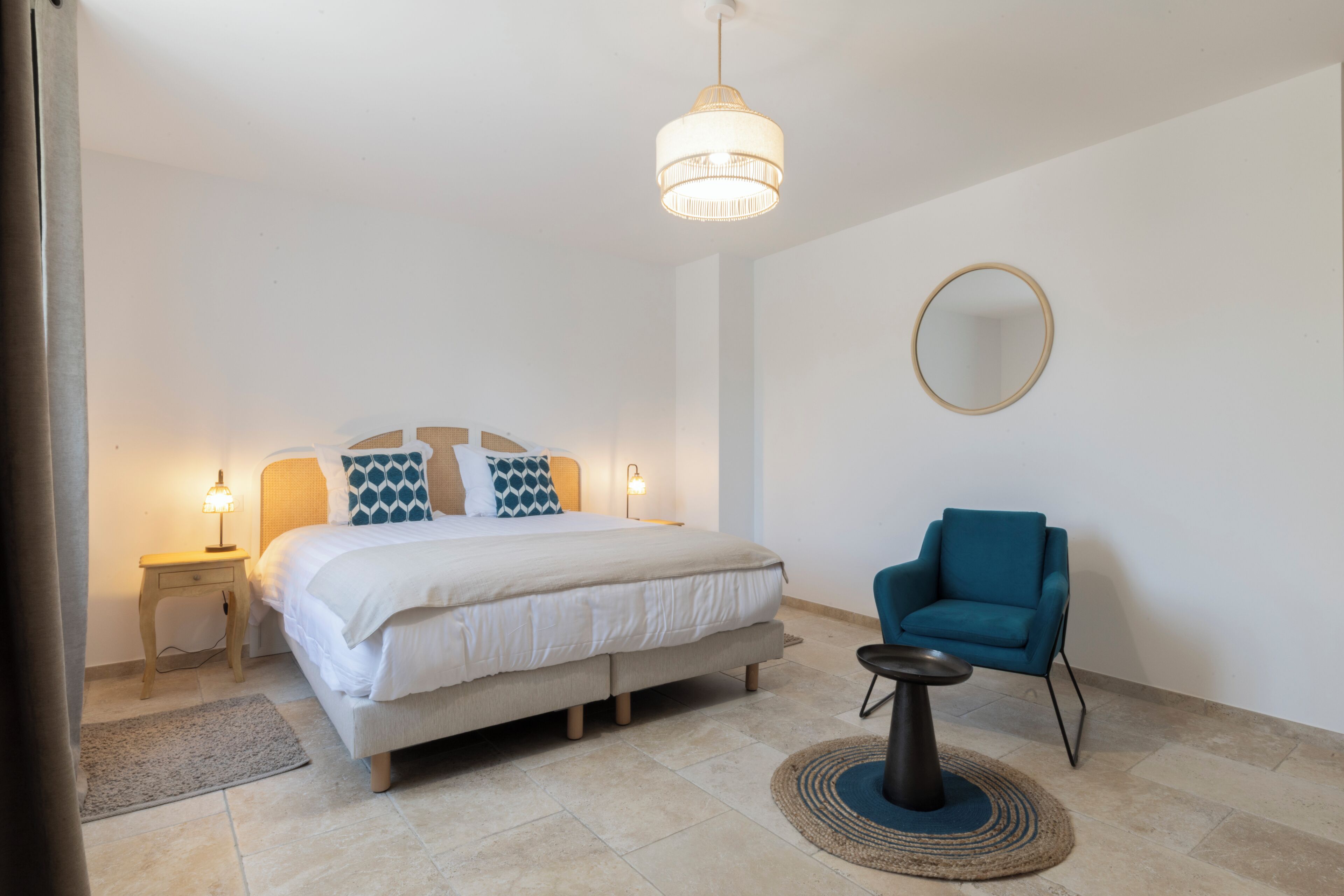 A bedroom with a bed, bedside tables, a chair, a round mirror, and a hanging light fixture.