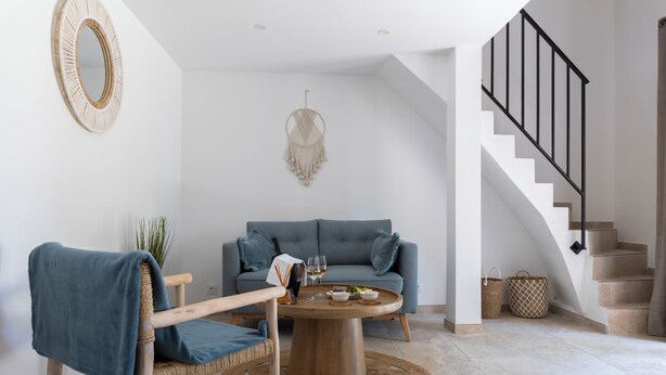 A modern living room with a round wooden coffee table, a blue sofa, a wicker chair, and a staircase with a black railing.