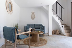 Una sala moderna con una mesa de centro redonda de madera, un sofá azul, una silla de mimbre y una escalera con barandilla negra.