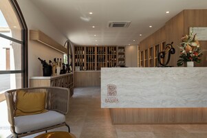 Una zona de recepción con mostrador de mármol, estanterías de madera con botellas, una silla de mimbre y un ventanal grande.